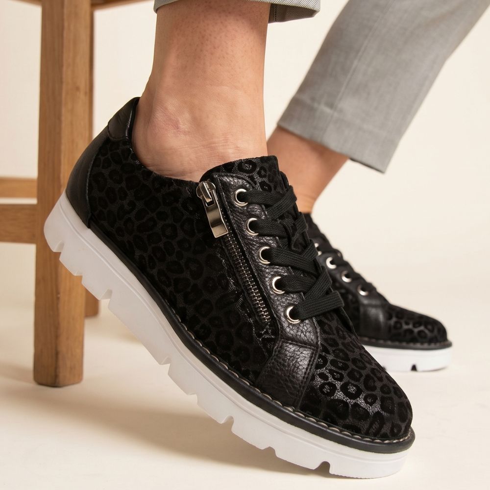 Black leopard print sneakers with white soles worn by a person sitting on a wooden chair.