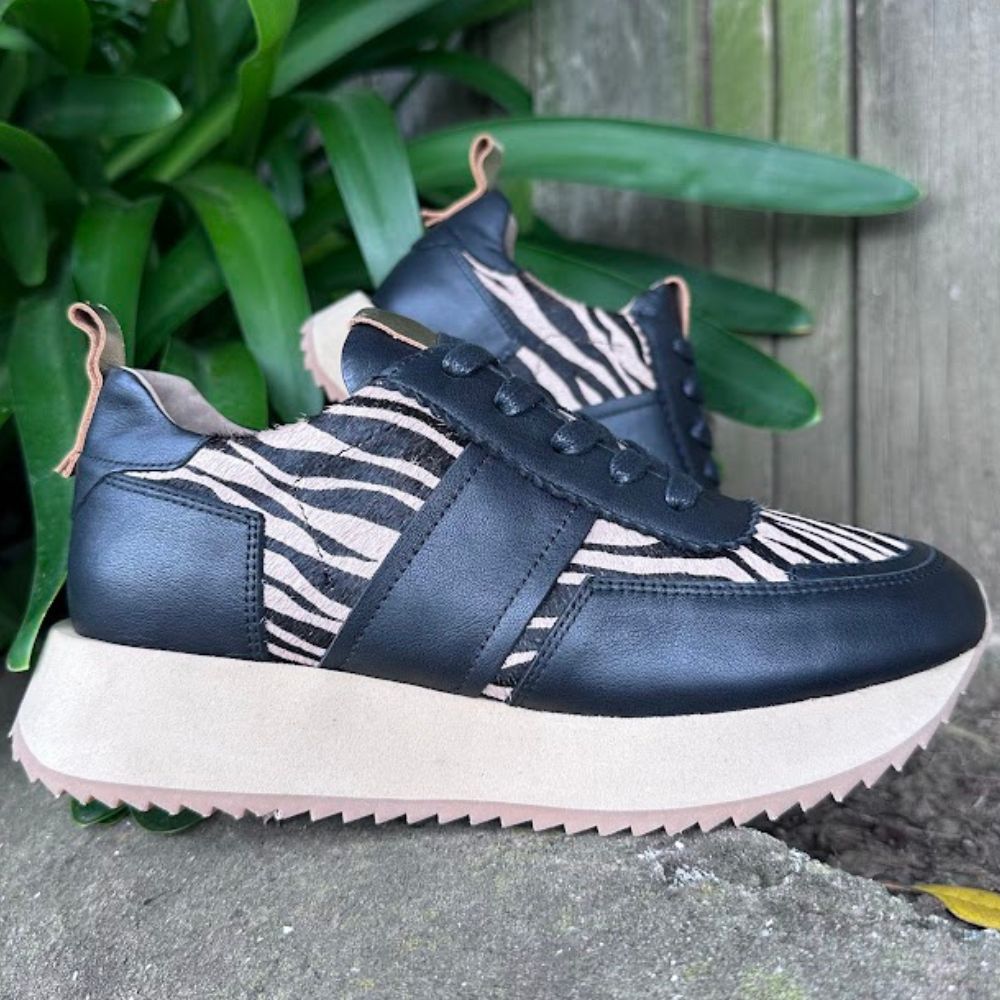 Black and tan zebra-patterned sneakers on a concrete surface with green plants in the background