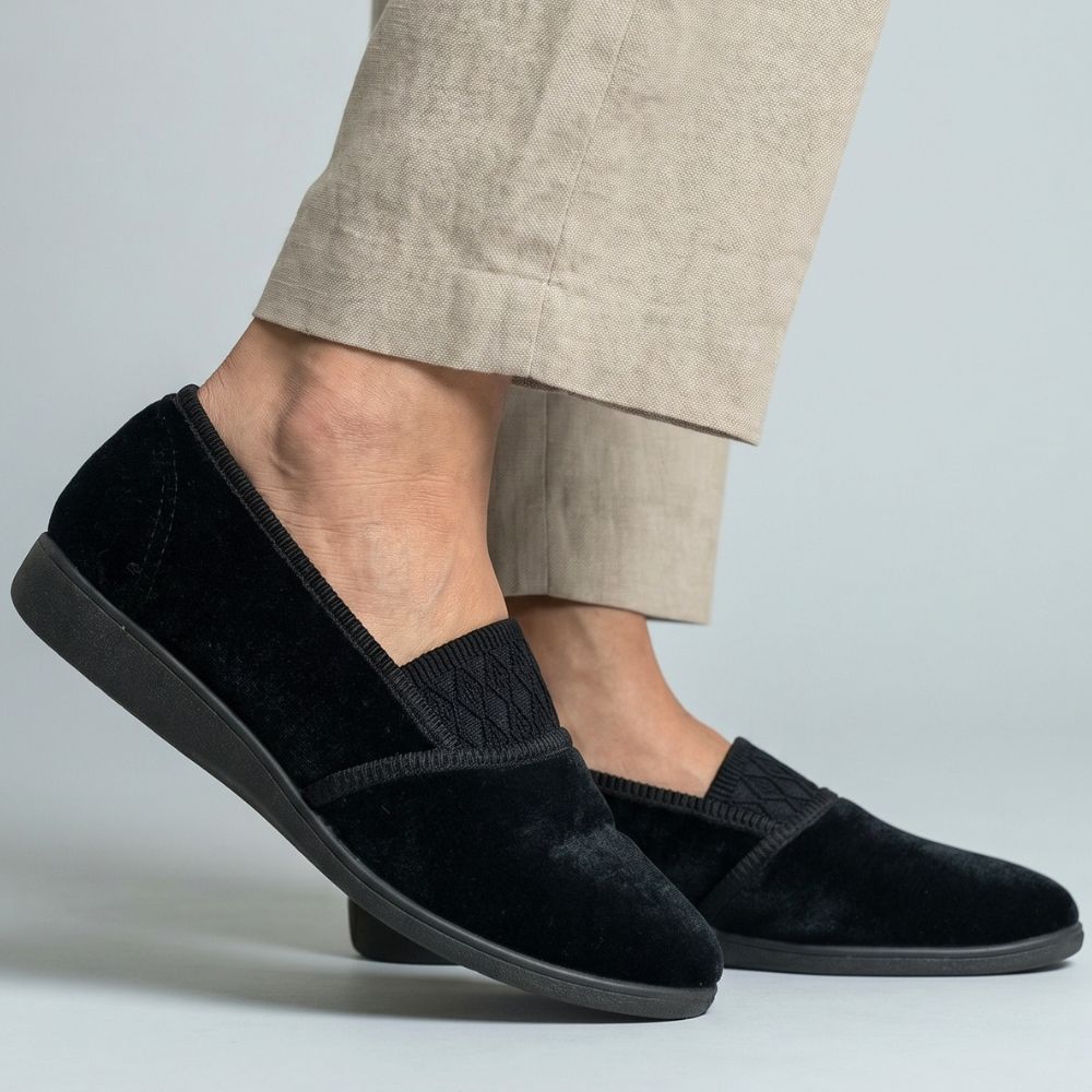Black slip-on slippers worn with beige pants on a light gray background