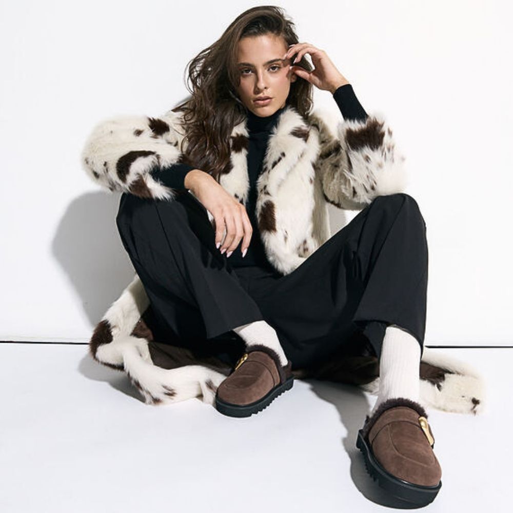 Woman wearing a cowhide coat and brown suede mules with fur lining on a white background