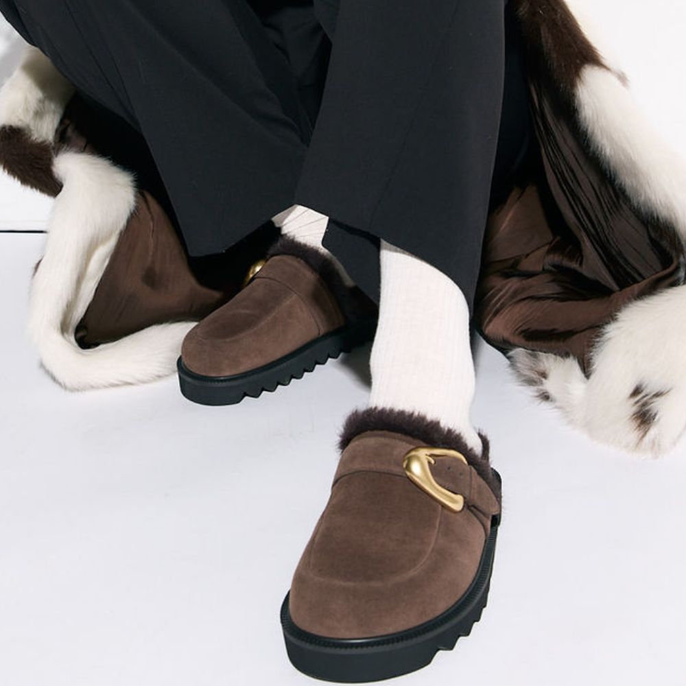 Brown suede mules with fur lining and gold buckles worn by a person sitting on a fur surface.