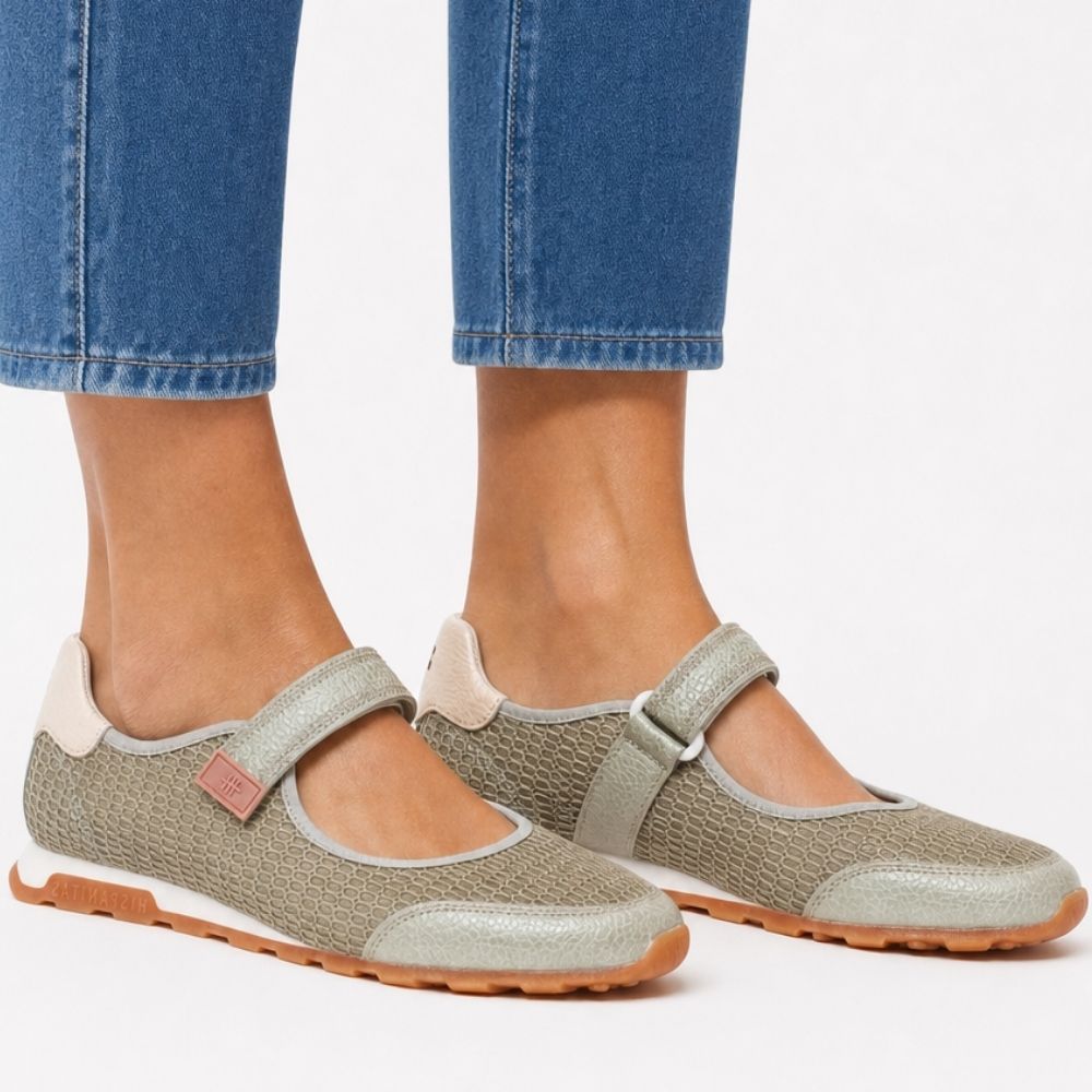A pair of women's Mary Jane shoes in a light sage textured mesh and leather, featuring hook-and-loop straps, cream heel counters, and tan rubber soles with the brand name 'Hispanitas', worn by a person wearing jeans on a white studio background