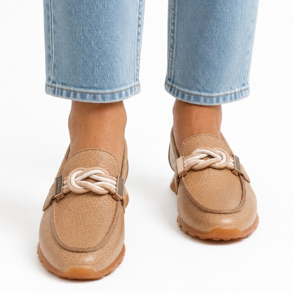 A pair of tan leather loafers with a tan and cream braided detail across the vamp, shown on a model's feet with light-wash blue jeans against a white background.