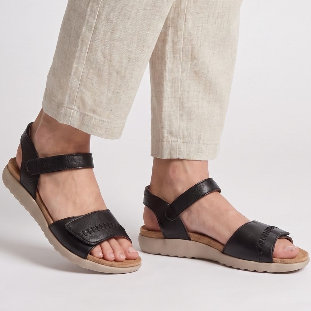 A pair of black leather slide sandals with adjustable straps and beige soles, shown on a model wearing light beige linen pants against a clean white studio background.