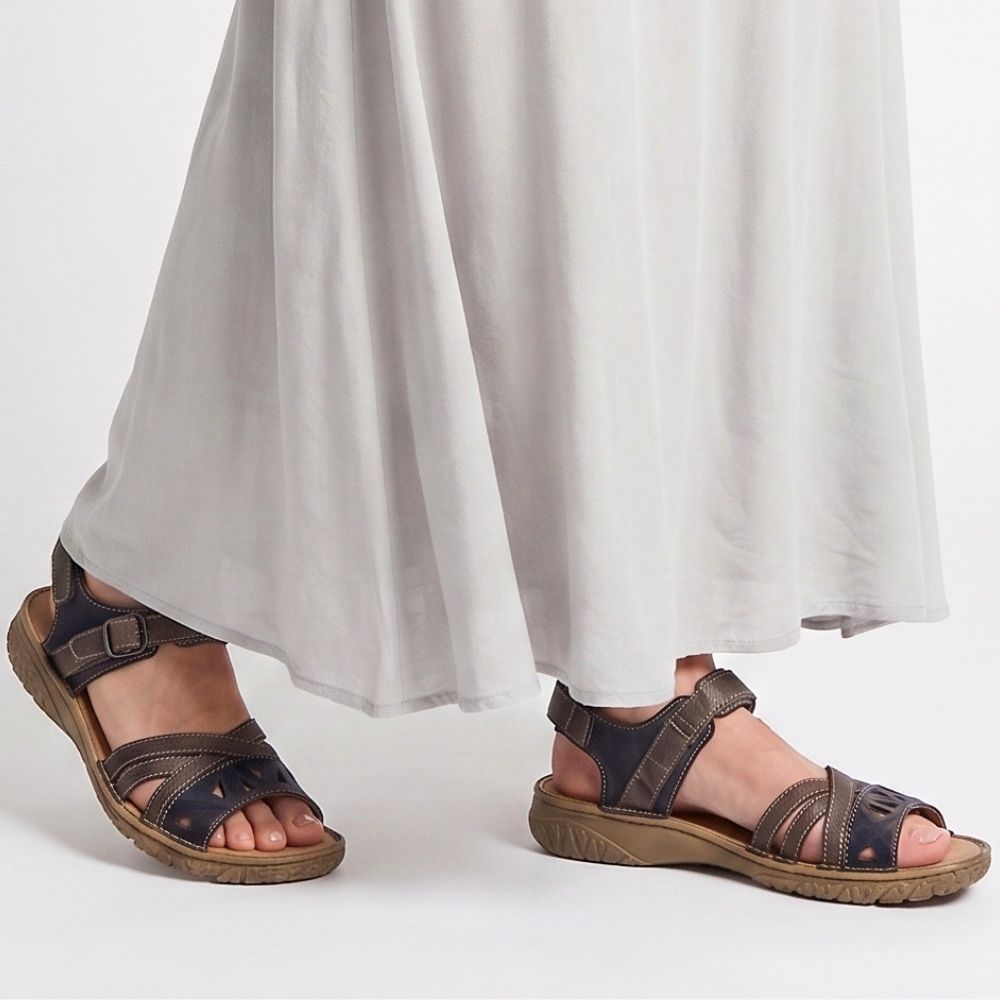 A pair of dark brown and navy blue leather strappy sandals with adjustable straps and thick brown soles, shown on a model wearing a flowing white maxi skirt against a plain white studio background.