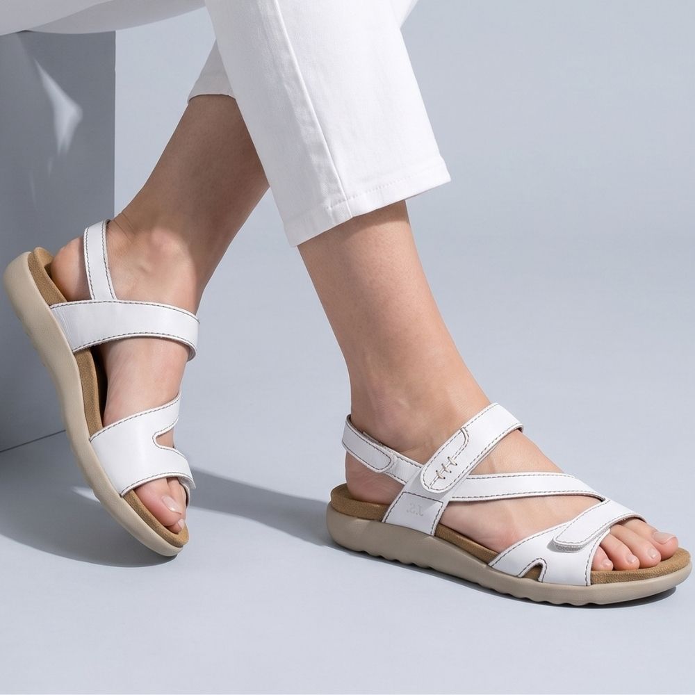 White leather strappy sandals with a tan footbed and sole, shown on a model's feet wearing white cropped pants against a light gray studio background.