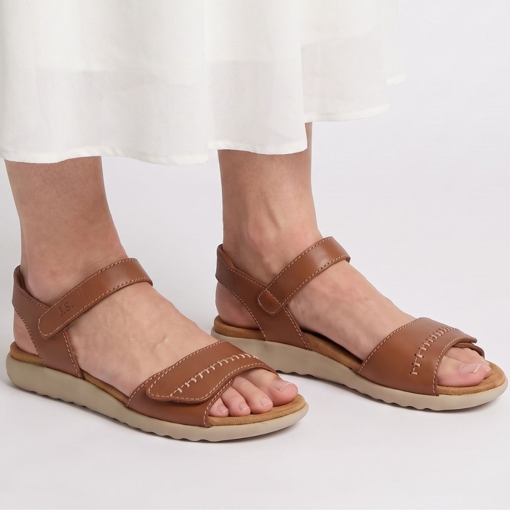 A pair of women's tan leather slide sandals with adjustable straps and beige soles, shown on a model wearing a white skirt against a light gray studio background.