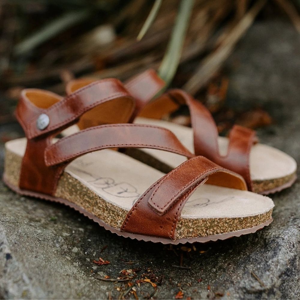 A pair of brown leather strappy sandals with cork-look midsoles, photographed outdoors on a stone surface with blurred natural background elements.