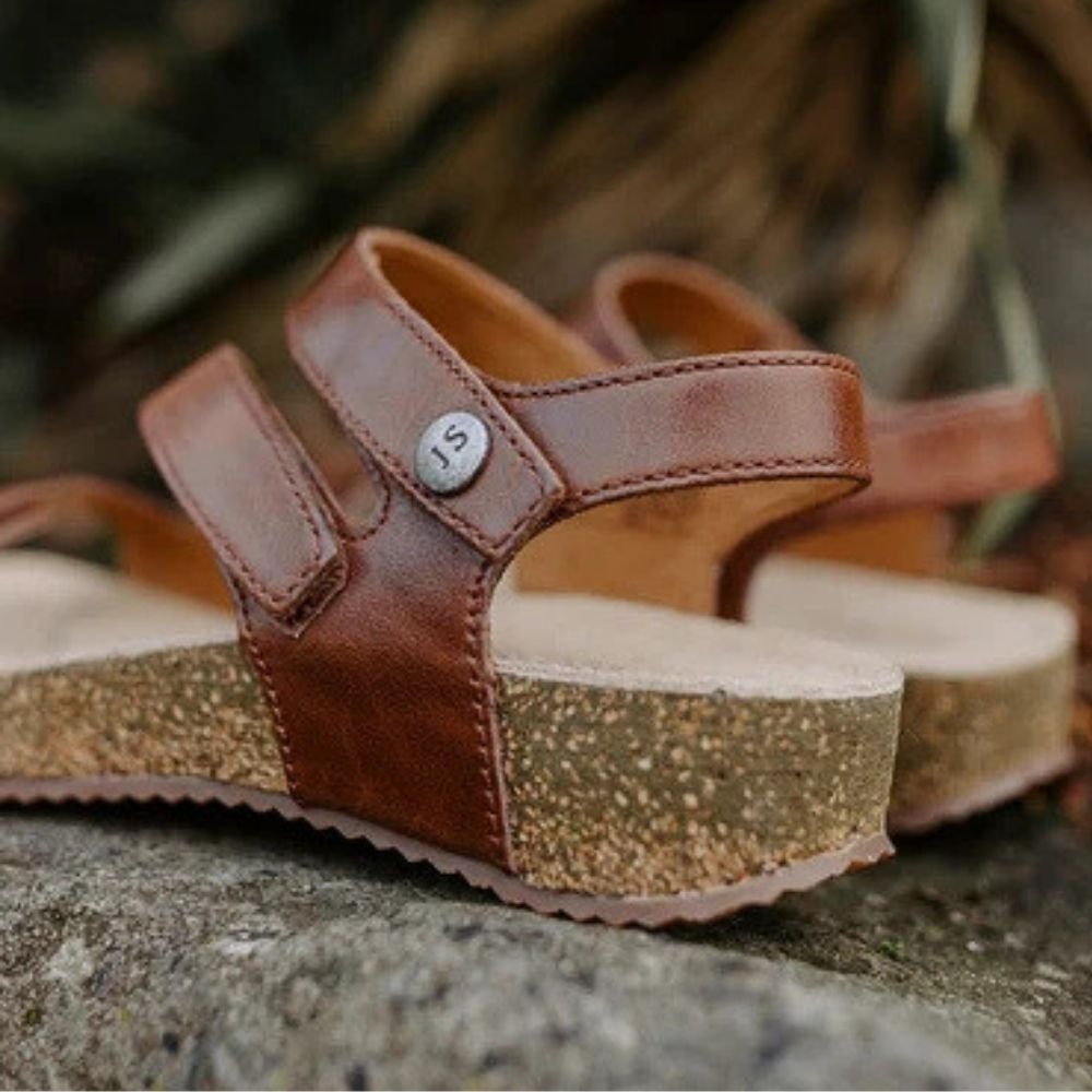 A pair of brown leather platform sandals with wide straps and a chunky cork-look wedge sole, photographed outdoors on a stone surface with a blurred natural background.