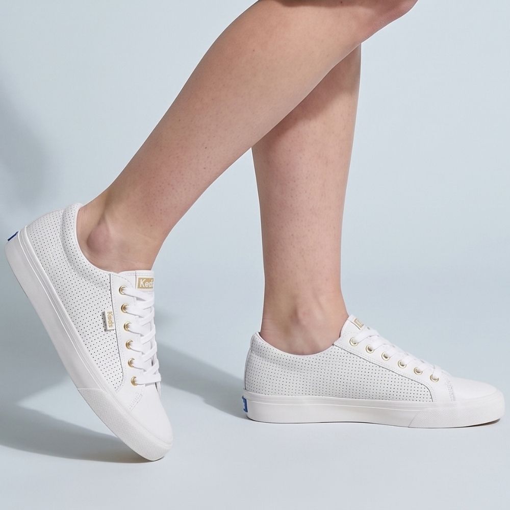 A person wearing white perforated leather KEDS JUMP KICK sneakers with gold embellishments and a lace-up design, against a blue studio background.