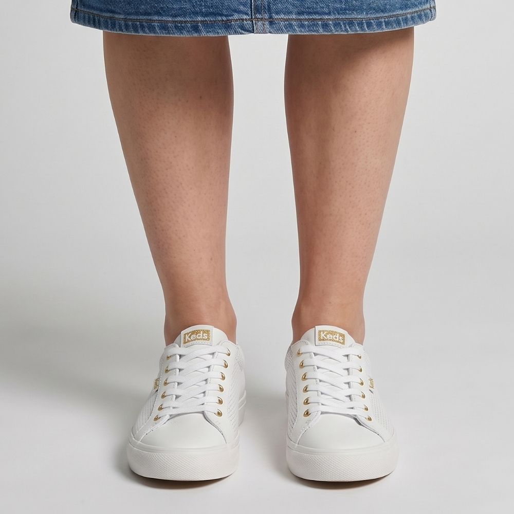 A person wearing a blue denim skirt and white perforated leather KEDS JUMP KICK sneakers with gold embellishments and a lace-up design, against a studio background.