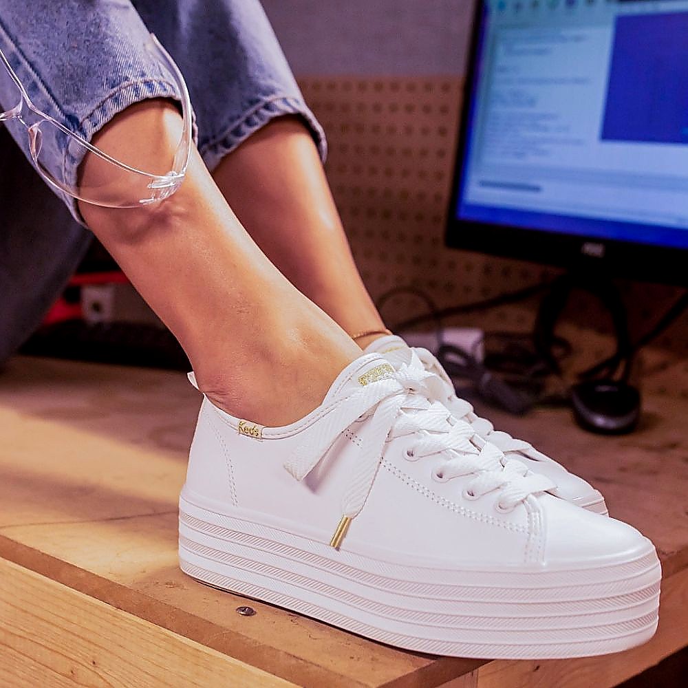 A person seated with their feet on a table wearing white KEDS TRIPLE UP LEATHER platform sneakers with a lace-up design.