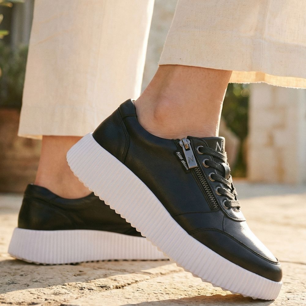 Black sneakers with white soles worn by a person on a stone surface.