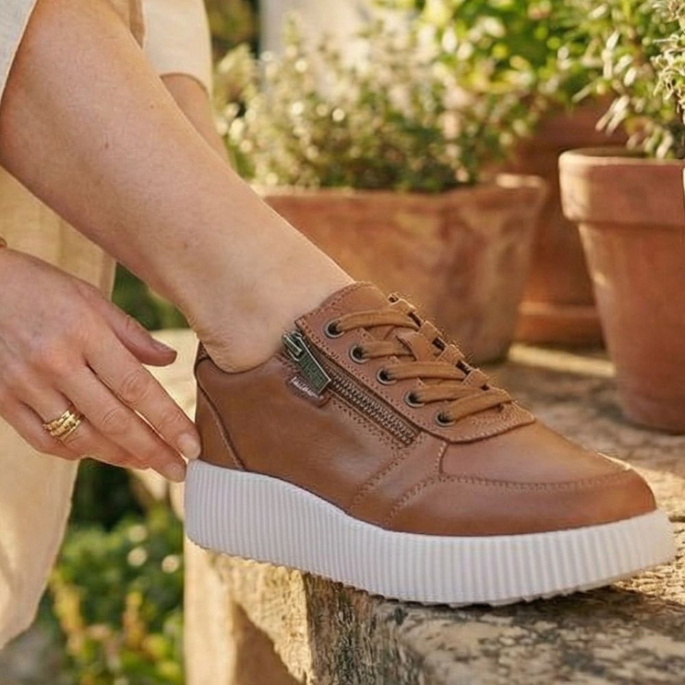 Tan sneaker with white sole worn by a person, with plants in the background