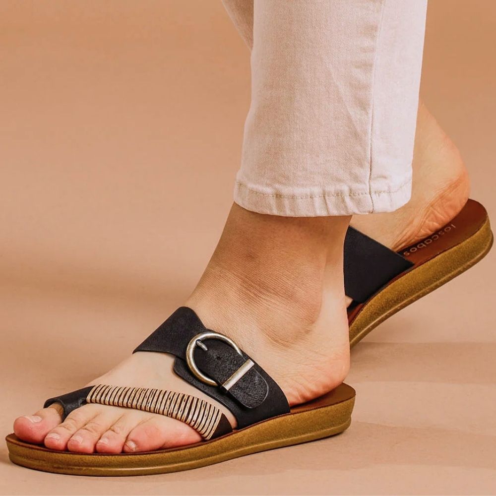 A close-up of a person's feet wearing black slide sandals with a braided bamboo toe strap and an adjustable gold buckle, paired with light beige pants against an orange background.