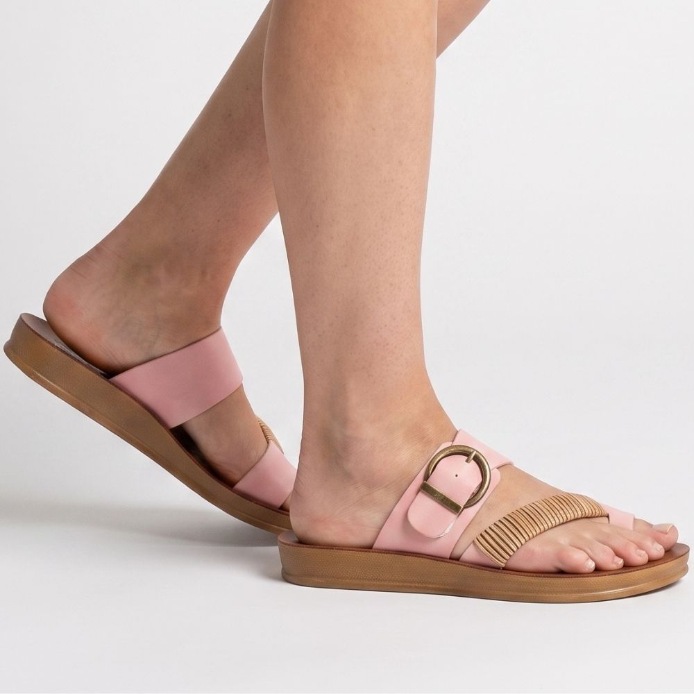 A woman wearing a pink slide sandal with an adjustable bronze buckle and braided rattan detail on a white background.