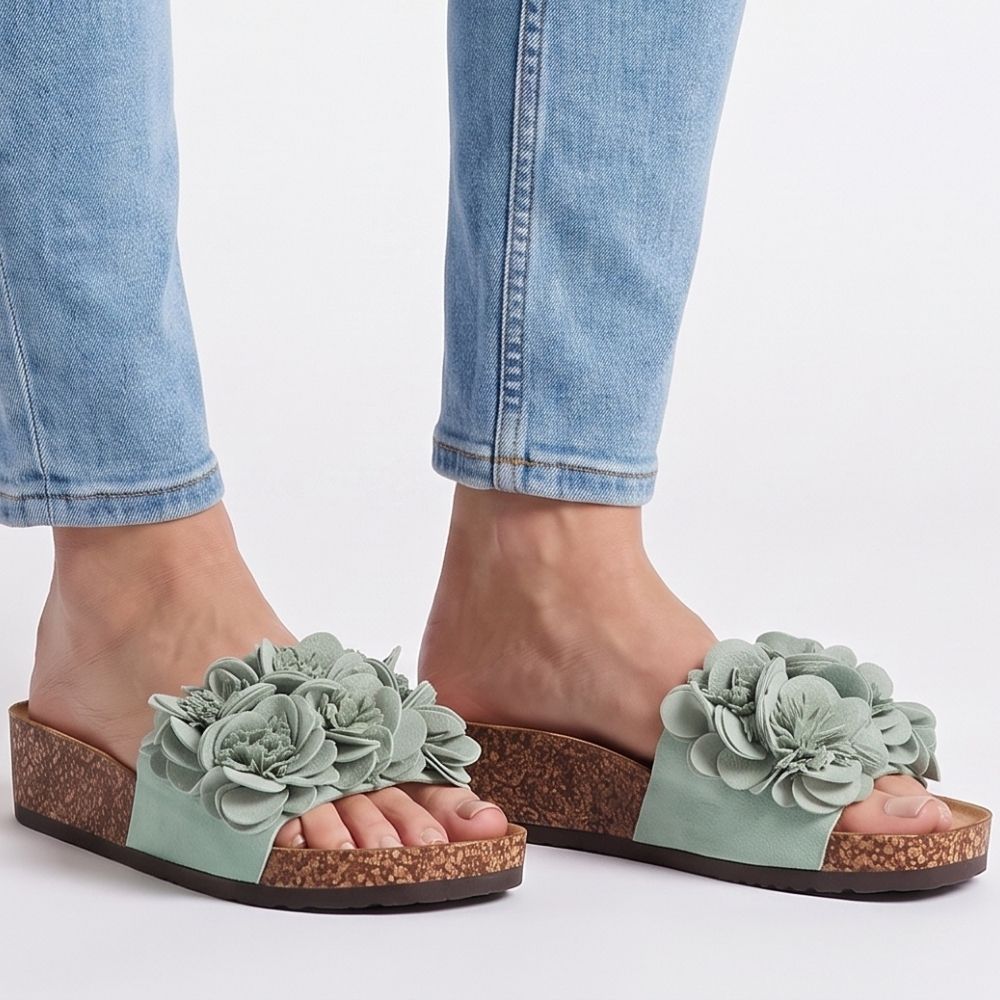 Person wearing blue jeans and mint slide cork wedge sandals with textured floral embellishments across the vamp, against a white background.