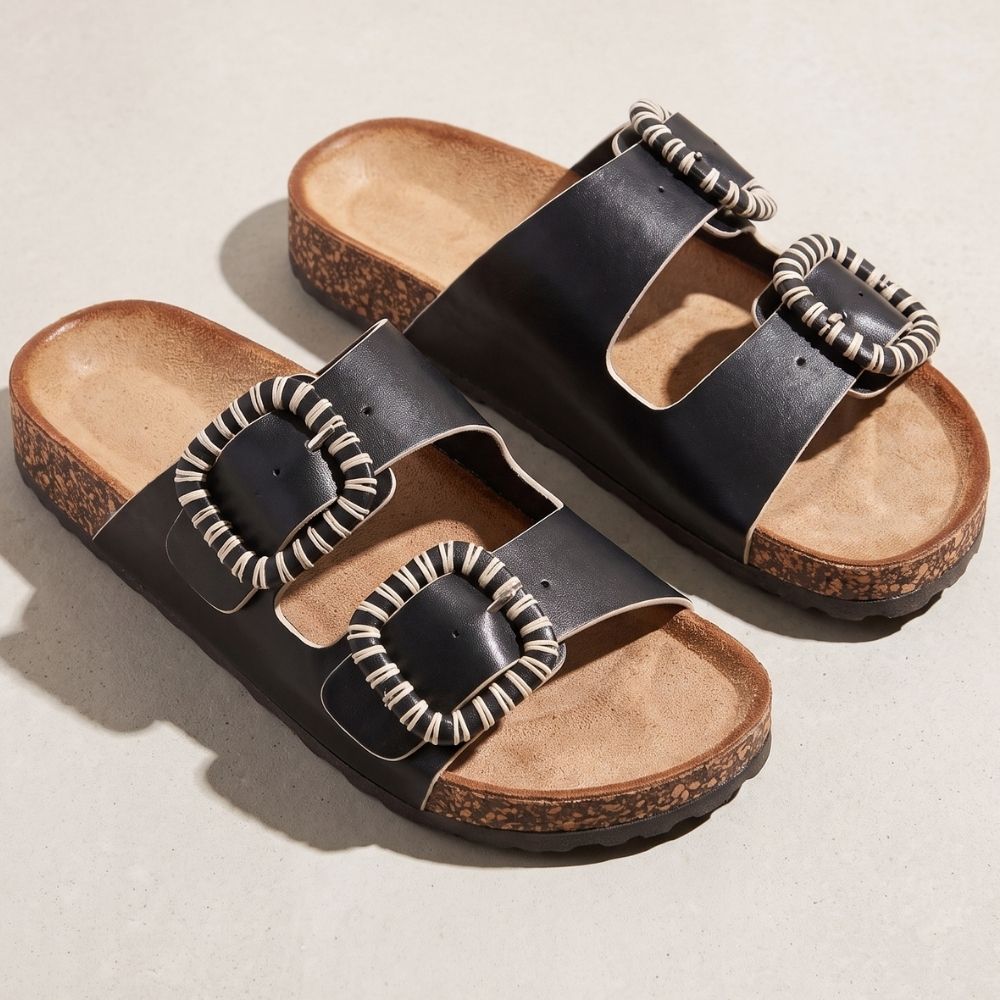 A pair of black double-strap slide sandals with textured woven trim around large square buckles, cork midsoles, and tan footbeds, photographed on a light neutral surface.