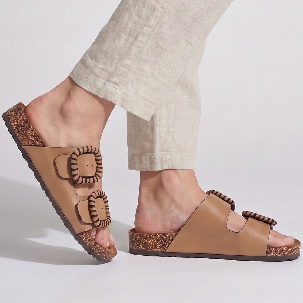 A person wearing linen pants and a pair of Vegan leather double-strap slide sandals with decorative woven trims around the straps and textured cork midsoles, shown against a plain white background.