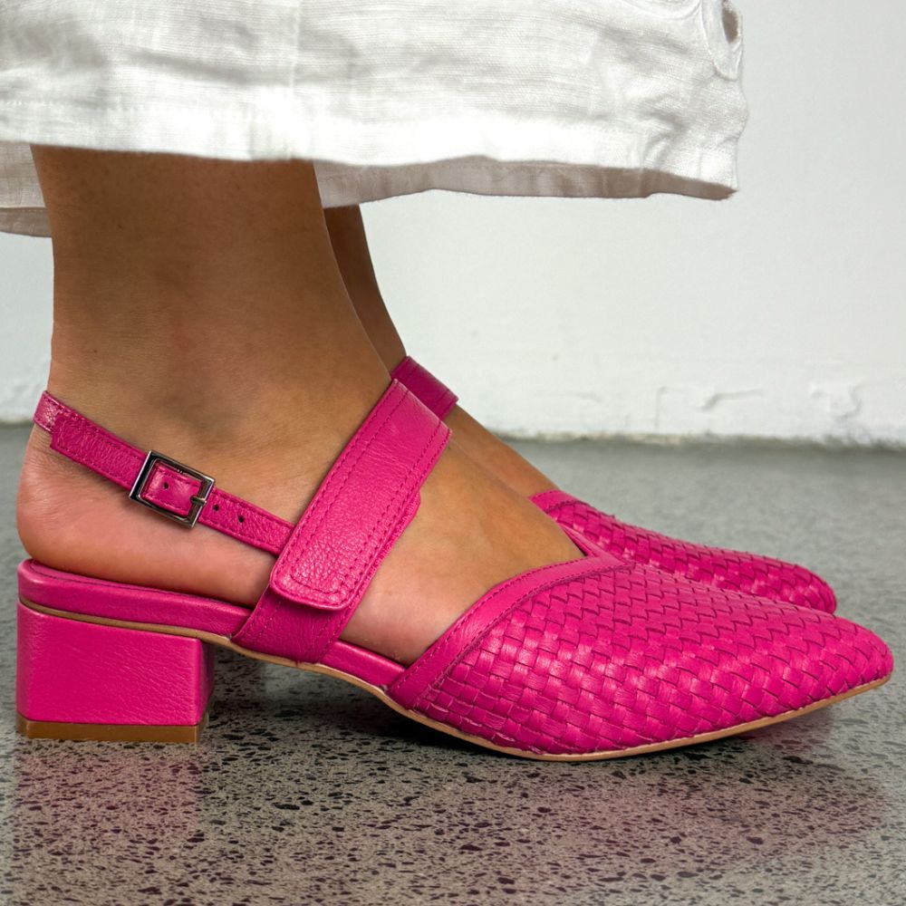 Bright pink woven leather heeled sandals with ankle straps and block heels, shown on a model's feet against a neutral indoor background.