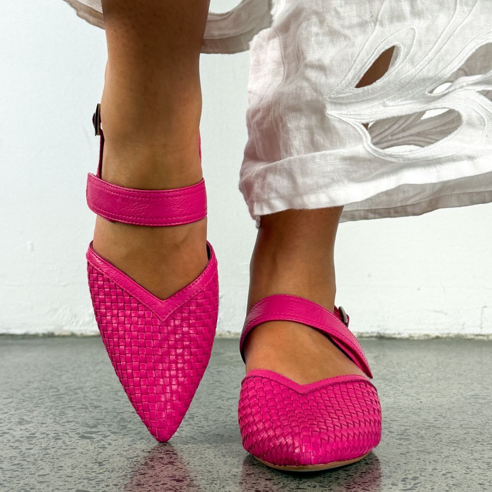 Bright pink woven pointed-toe heeled sandals with ankle straps, shown on a model wearing a white skirt against a light wall and gray floor.