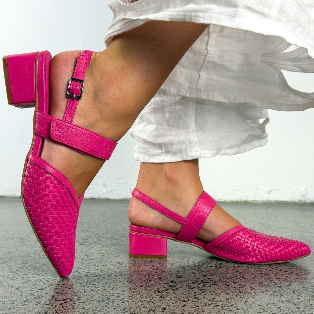 A person wearing bright pink woven slingback heeled sandals, shown against a white wall and gray floor.