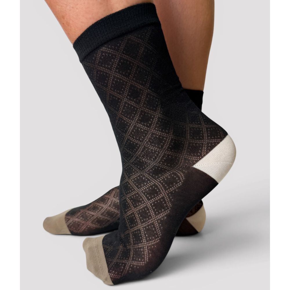 A pair of black crew socks with a subtle diamond lattice pattern and contrasting taupe toes and white heels, shown on a model's feet against a light gray background.