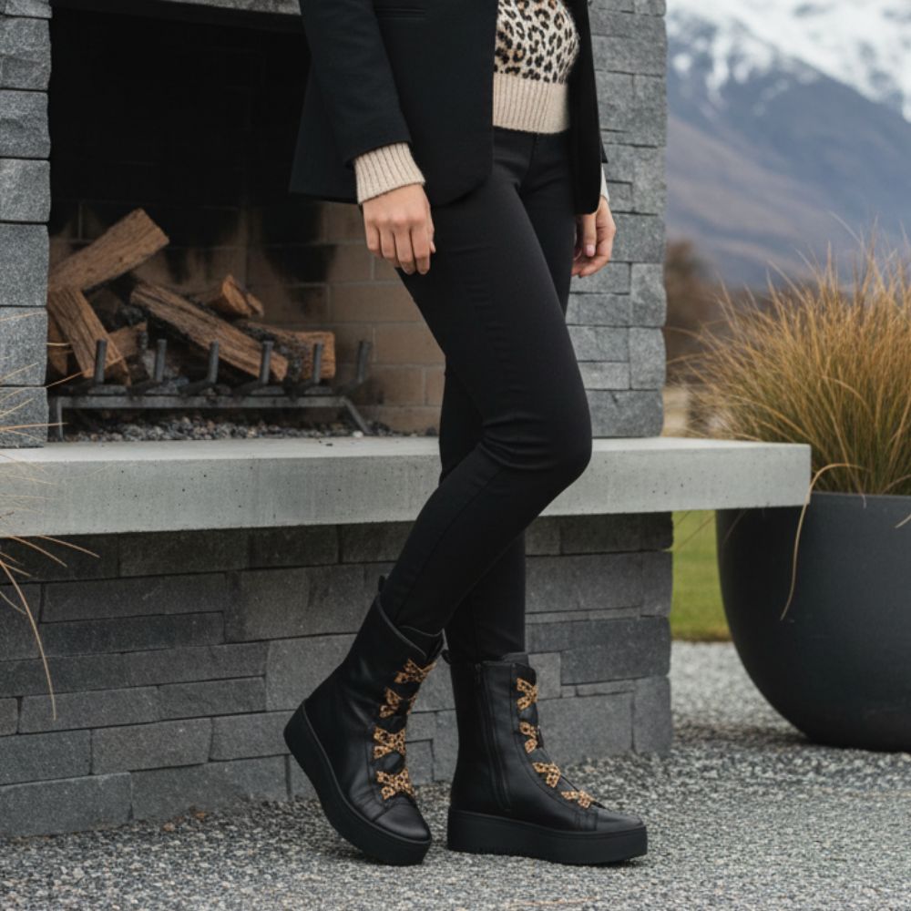 Person wearing black boots with leopard print patterns standing in front of a fireplace with mountains in the background.