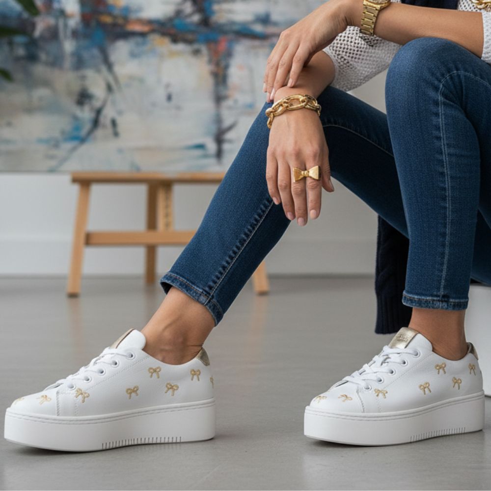Person wearing white sneakers with gold bow details, sitting on a light gray floor.