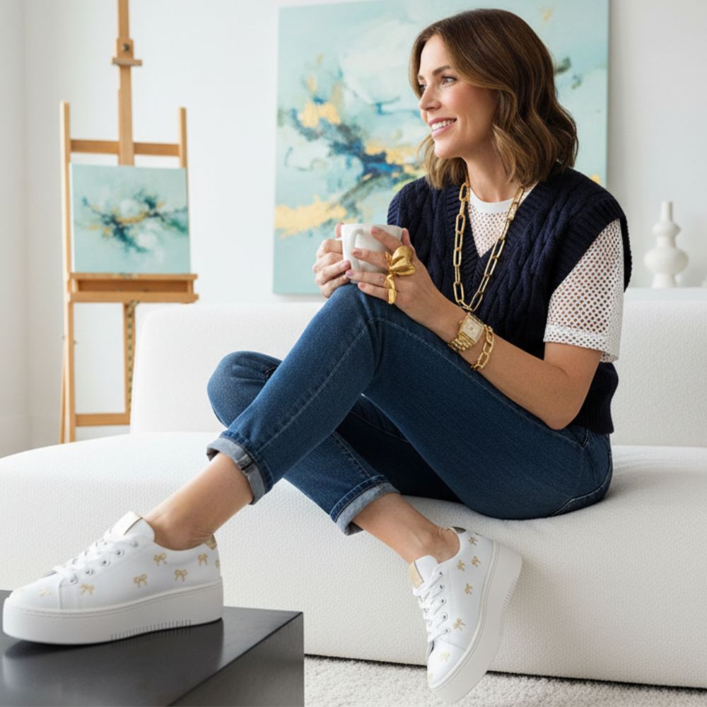 Woman sitting on a white stool wearing white sneakers with gold bows, holding a mug in a room with art and decor.