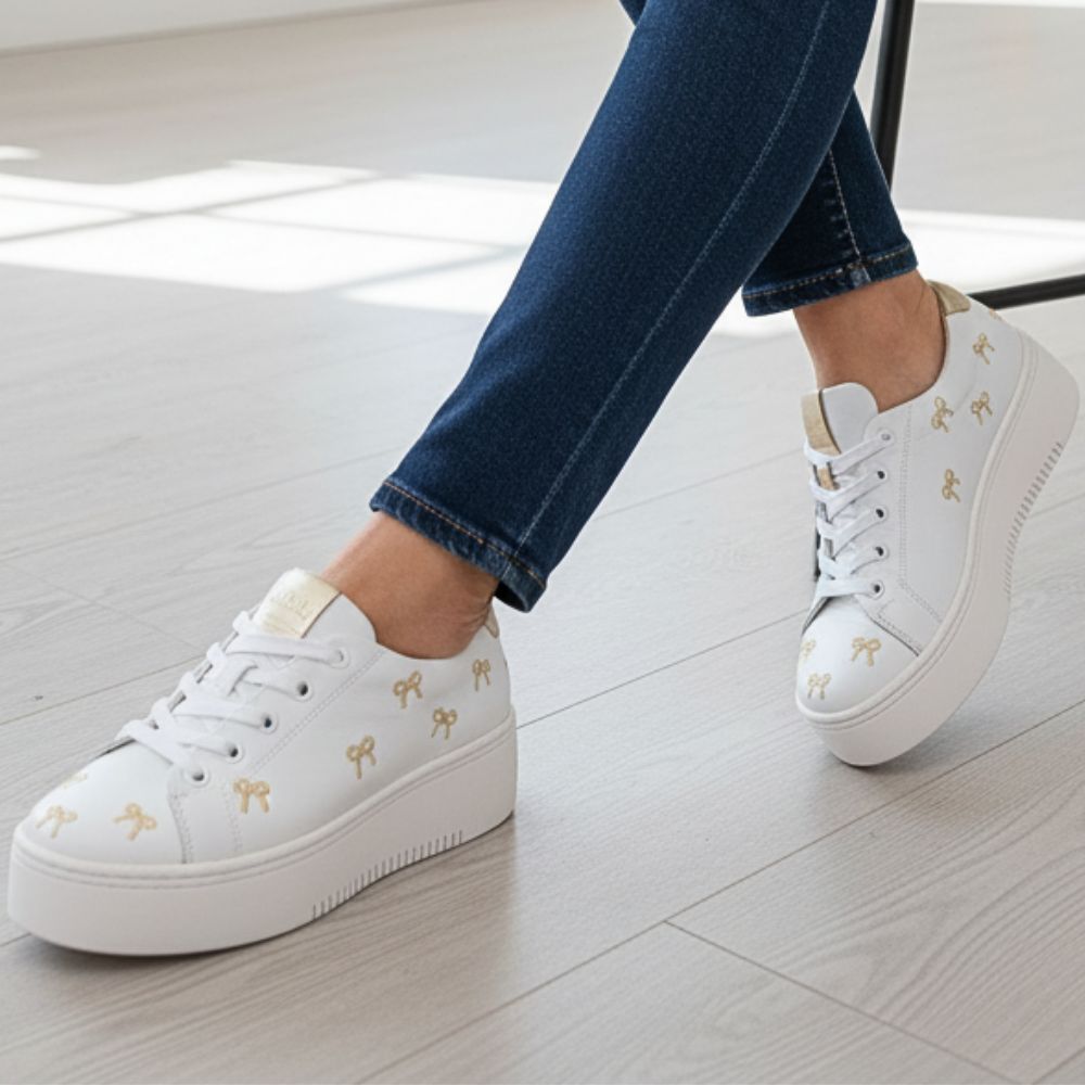 White sneakers with gold bows worn by a person on a light wooden floor.