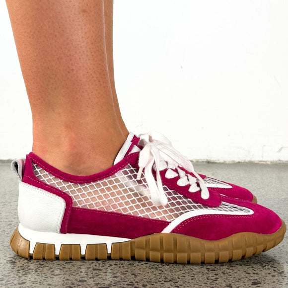 A person's foot wearing a pair of low-top sneakers with magenta suede, white mesh panels, white laces, and white heel accents, set against a white wall and speckled gray floor.