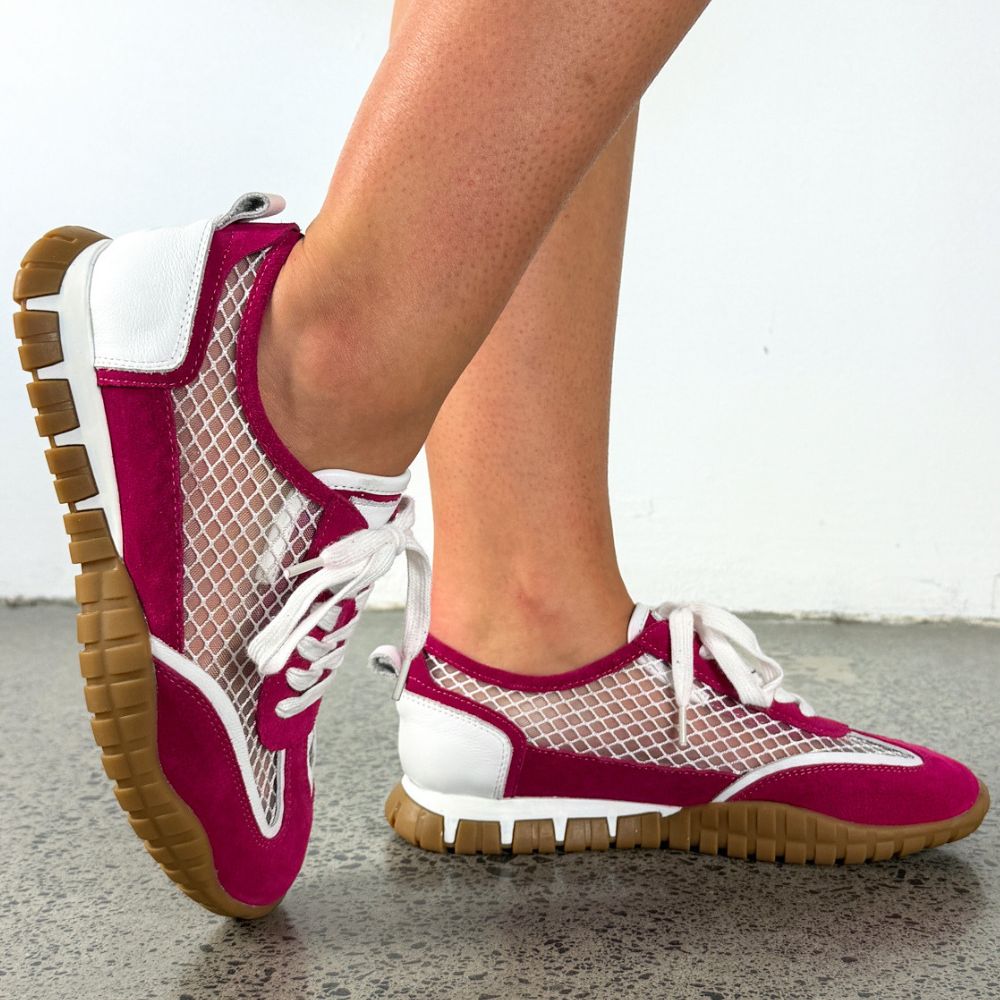 A pair of women's athletic sneakers worn on a person's feet, featuring a magenta pink and white mesh design with white laces and gum soles, set against a plain indoor background.