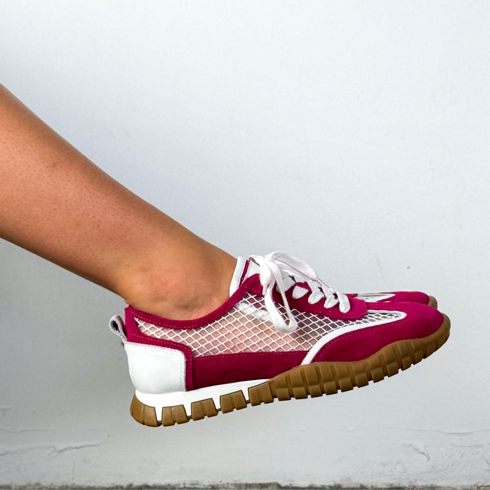 A close-up of a person's foot wearing a low-top sneaker with a magenta-pink upper, white mesh panels, white laces, and a brown gum sole, set against a plain light gray wall.