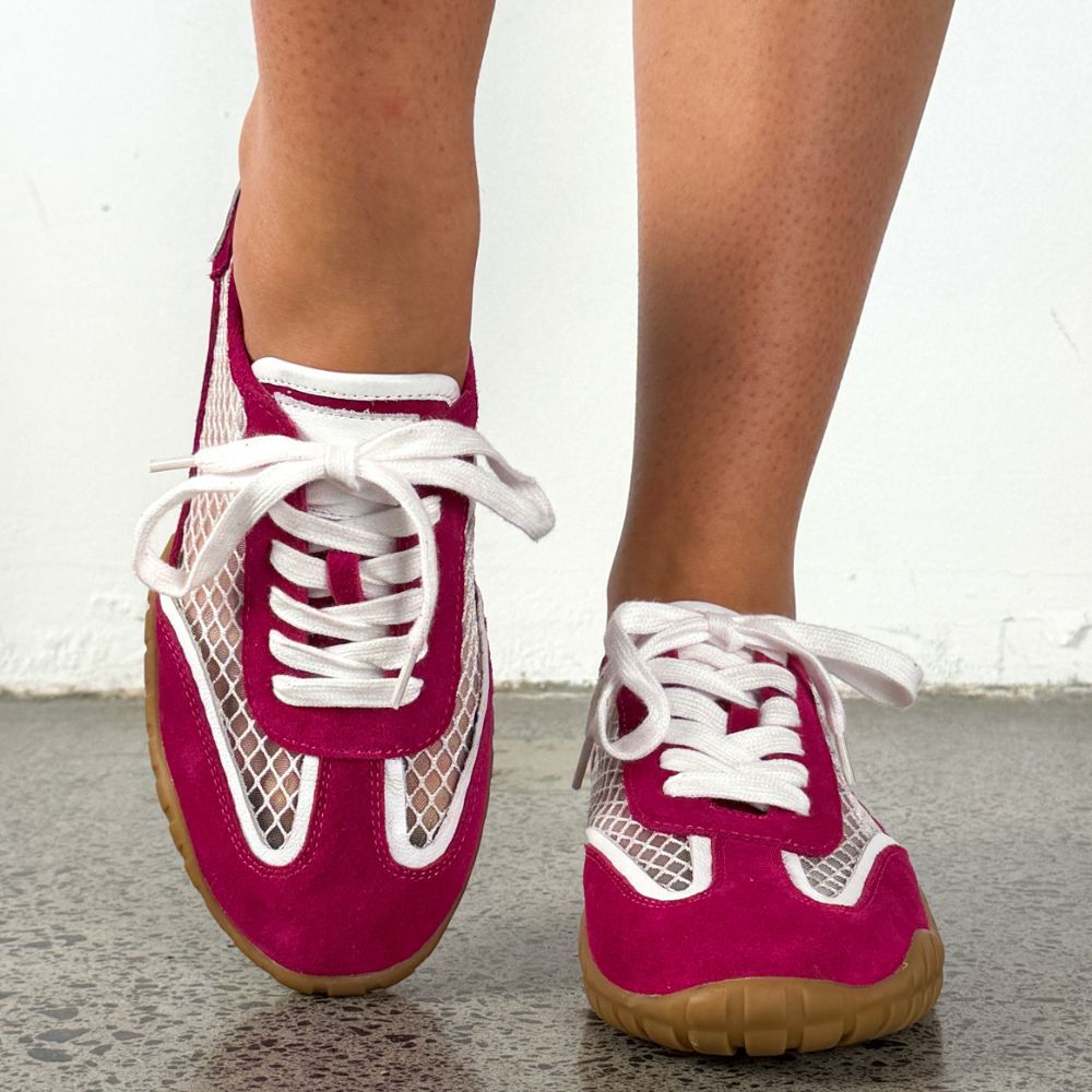 A close-up of a person's feet wearing low-top sneakers with bright magenta pink suede panels, white mesh side panels, white laces, and tan rubber soles, standing on a gray floor against a white wall.