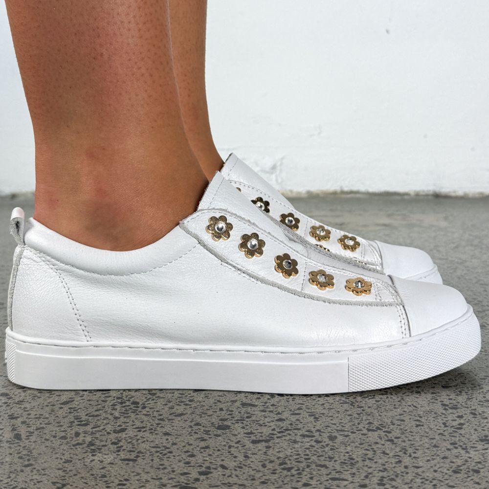 White leather slip-on sneakers with decorative gold-tone flower charms, shown on a person's feet against a gray concrete floor and white wall.