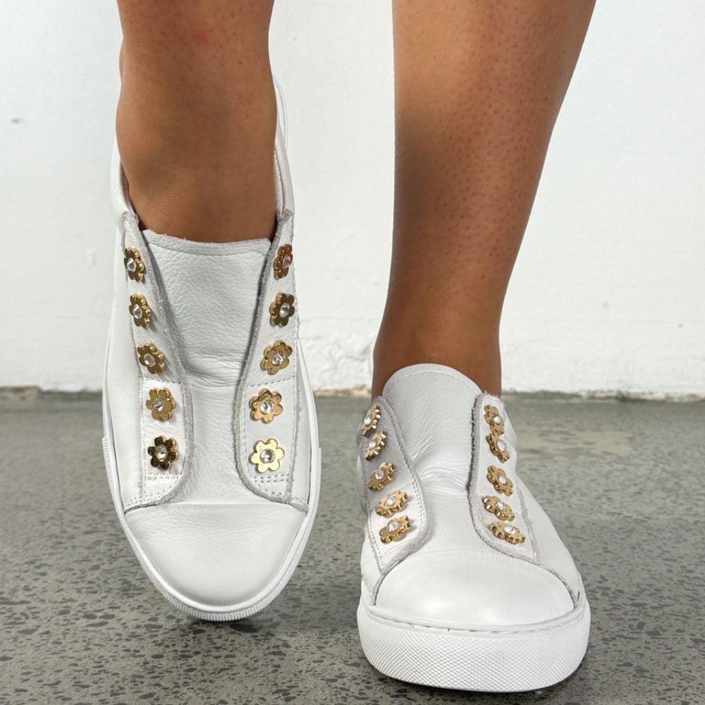 White leather slip-on sneakers with gold flower-shaped studs, shown on a person's feet against a white wall and gray floor.