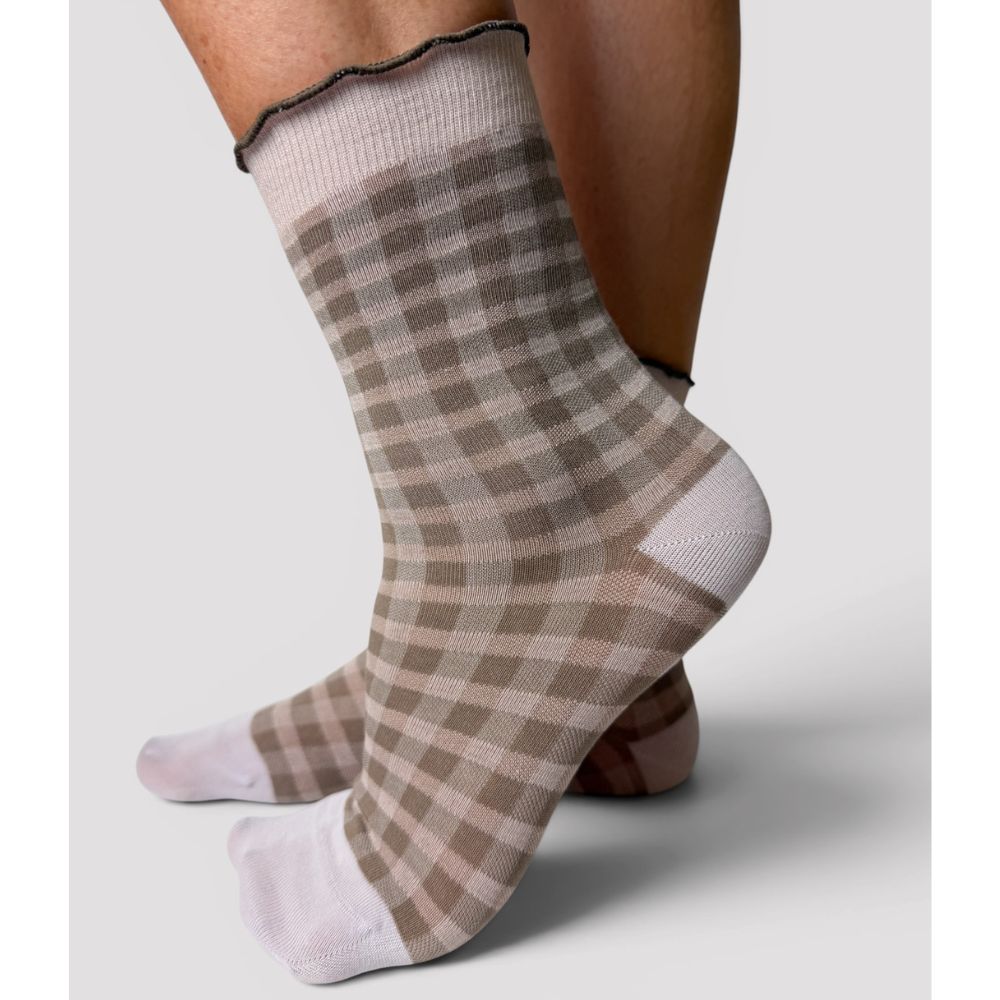 A pair of ankle socks with a beige, brown and pale pink plaid pattern and white toes and heel, shown on a person's feet against a light gray background.