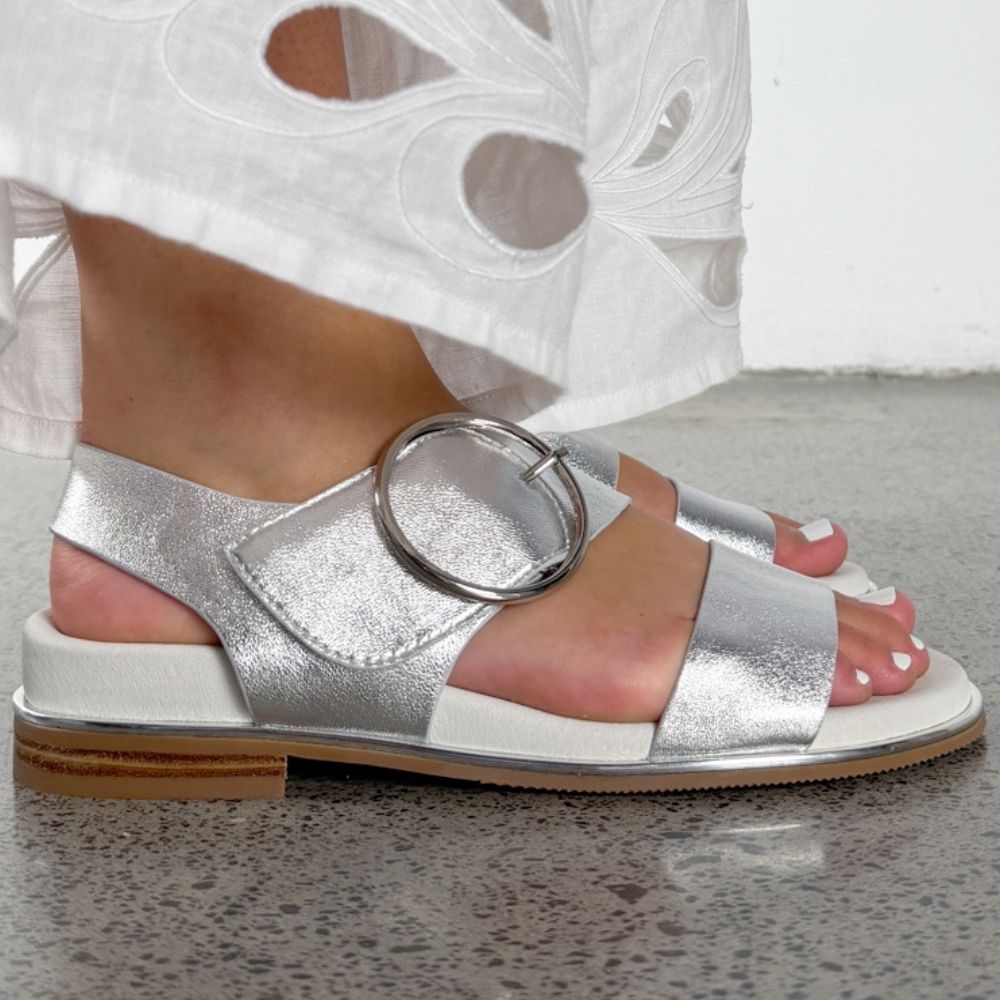 A close-up of a person wearing metallic silver flat sandals with a large circular buckle detail, paired with a white skirt, standing on a speckled gray floor.