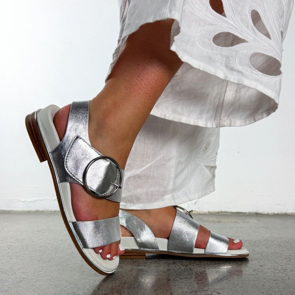 A person wearing metallic silver flat sandals with a large round buckle detail and white embroidered pants, shown from the knees down against a light gray wall and concrete floor.