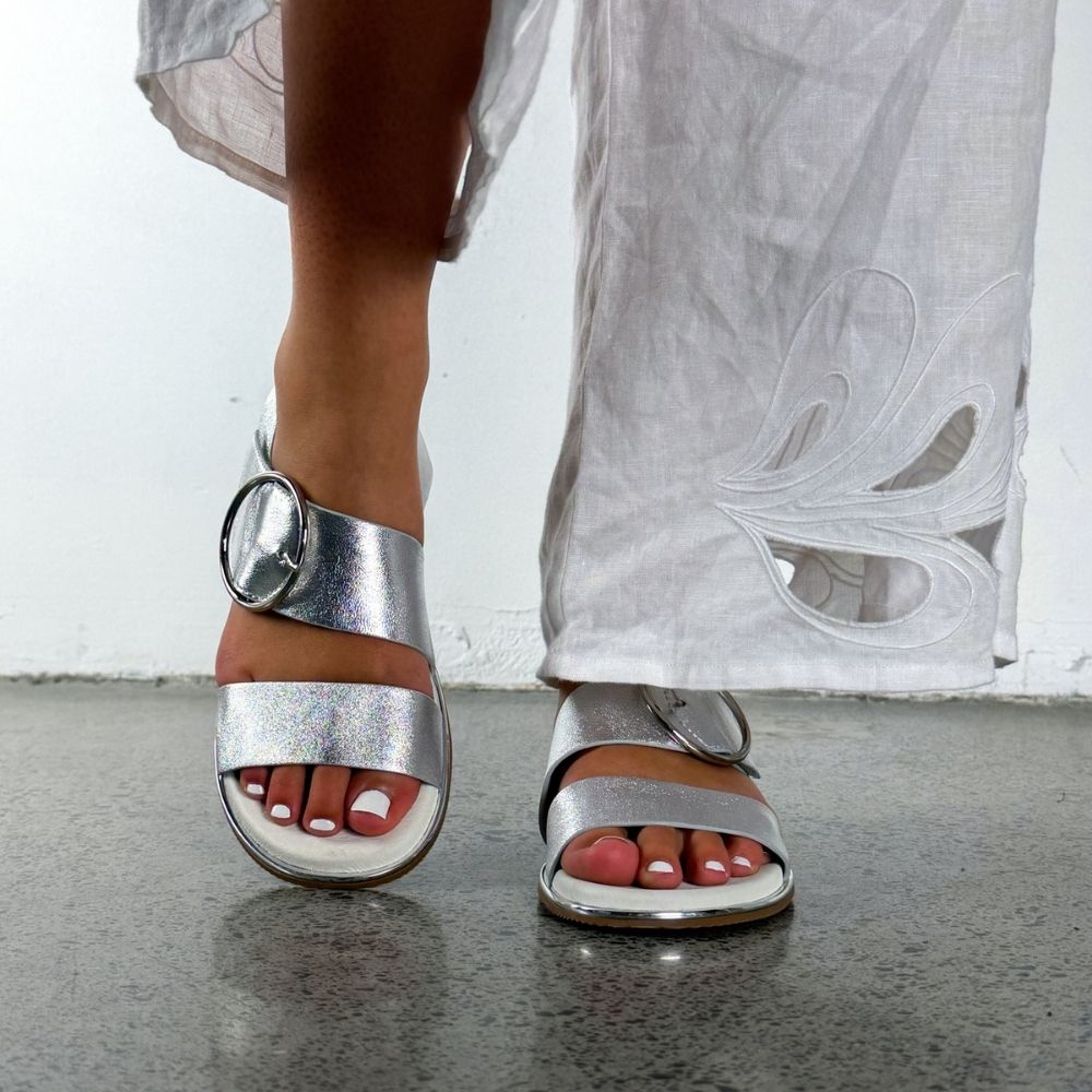 A close-up of a person wearing metallic silver strappy sandals with a large circular buckle detail, paired with a white skirt, standing on a gray floor.