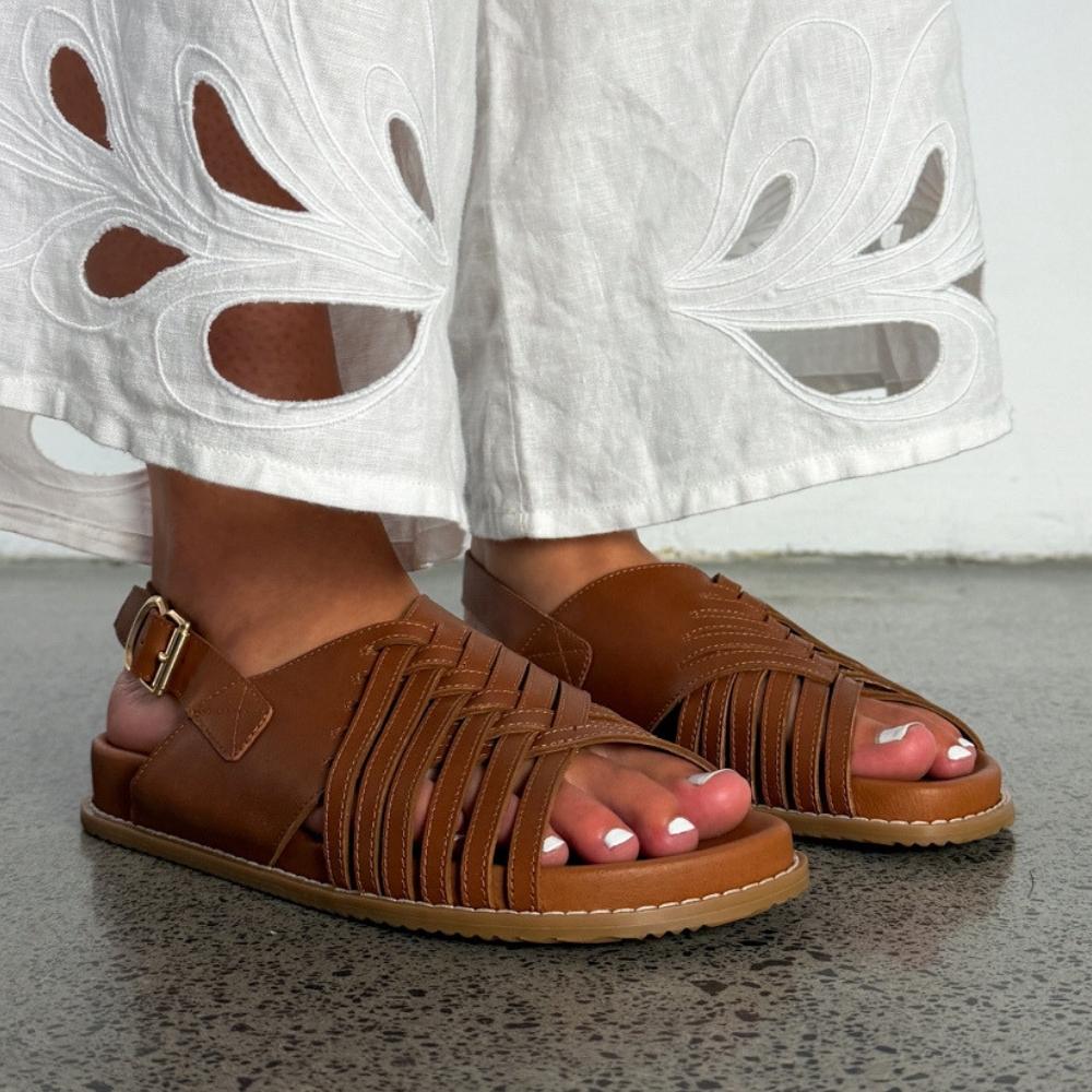 A pair of brown leather strappy flat sandals worn with white embroidered pants, shown on a person's feet against a gray floor and white wall.