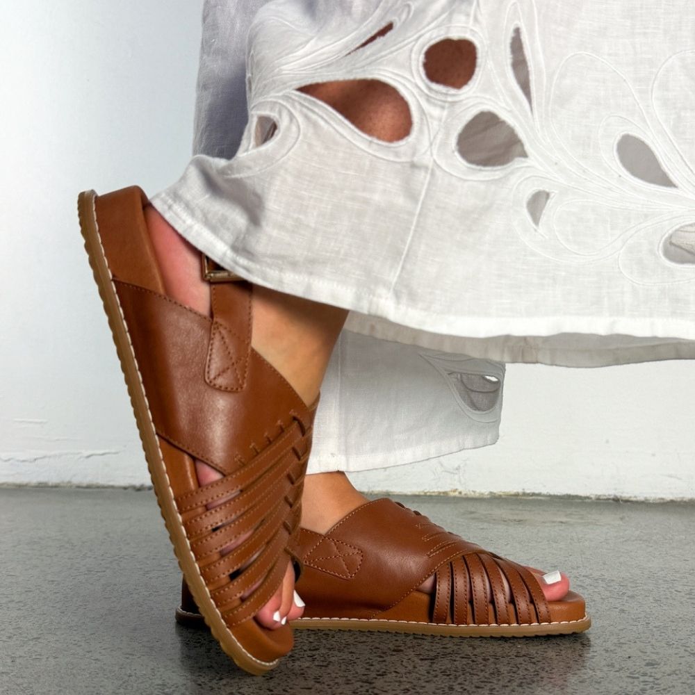 A person wearing brown leather strappy slide sandals with a slingback design, paired with a white embroidered skirt, standing on a gray floor against a white wall.