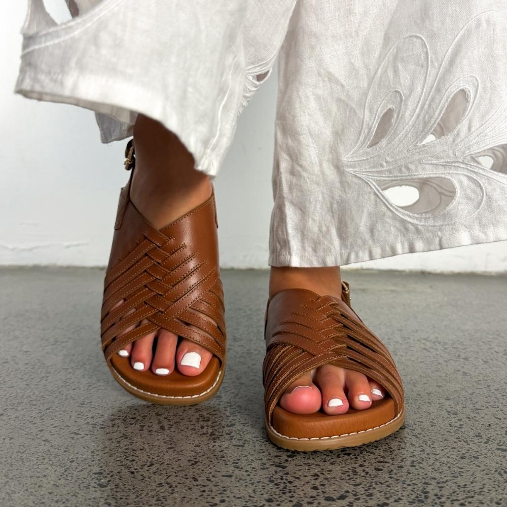 A pair of brown leather strappy sandals worn with white embroidered pants, shown on a person's feet against a gray floor and white wall.