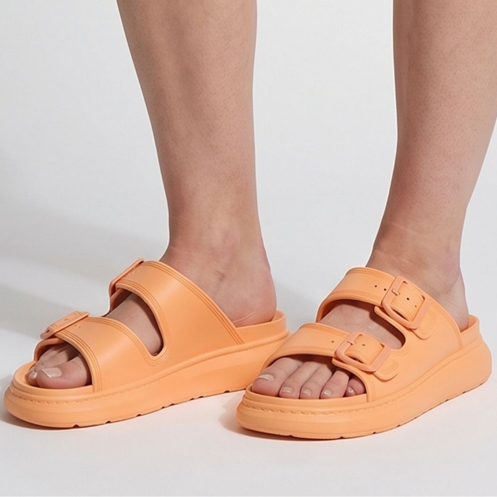 A person wearing a pair of orange platform rubber slide sandals with two adjustable buckles, against a studio background.