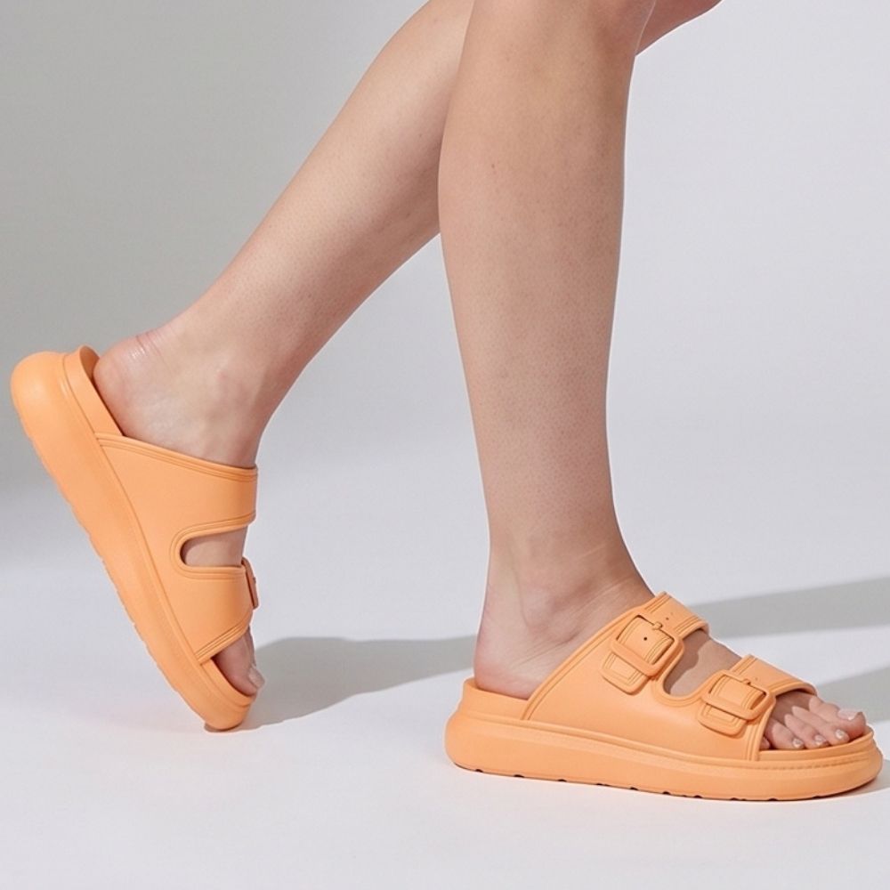 A person wearing a pair of orange platform rubber slide sandals with two adjustable buckles, against a studio background.
