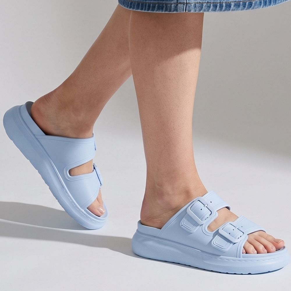 A person wearing a pair of sky-blue platform rubber slide sandals with two adjustable buckles, against a studio background.