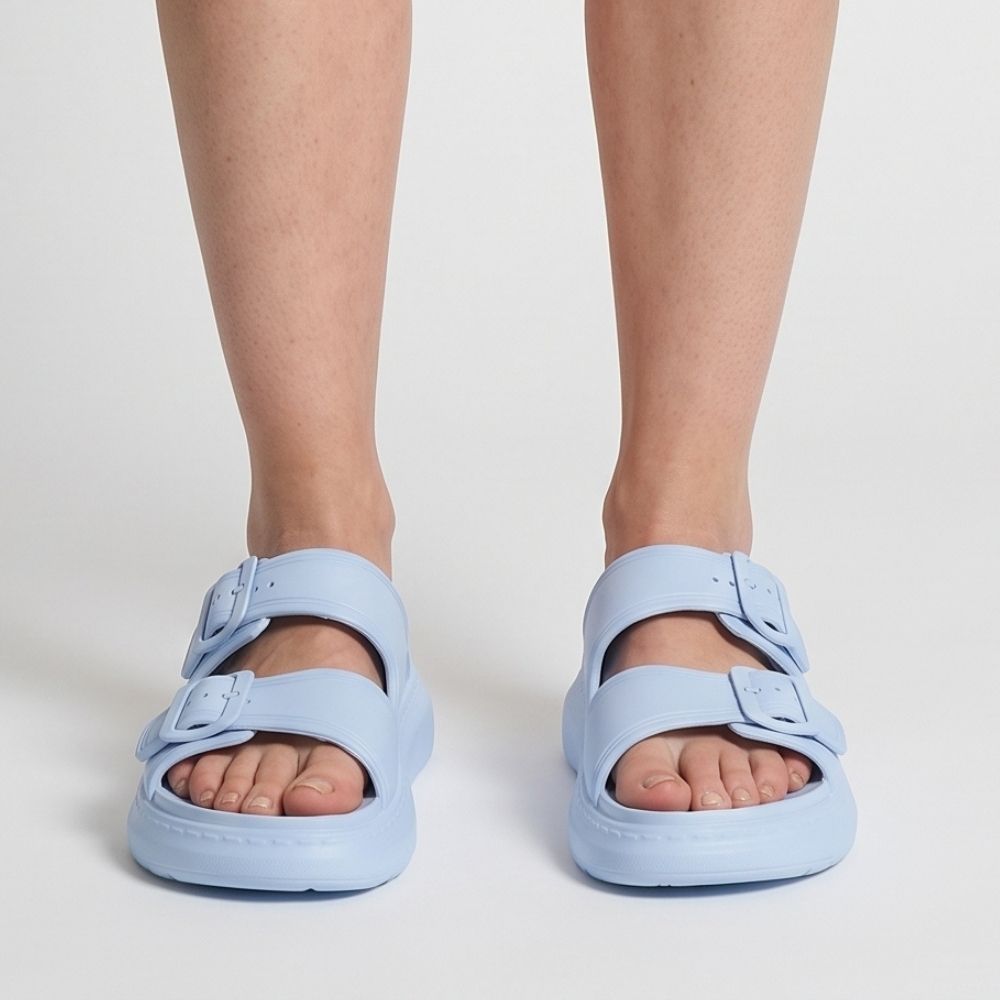 A person wearing a pair of sky-blue platform rubber slide sandals with two adjustable buckles, standing front-on against a studio background.