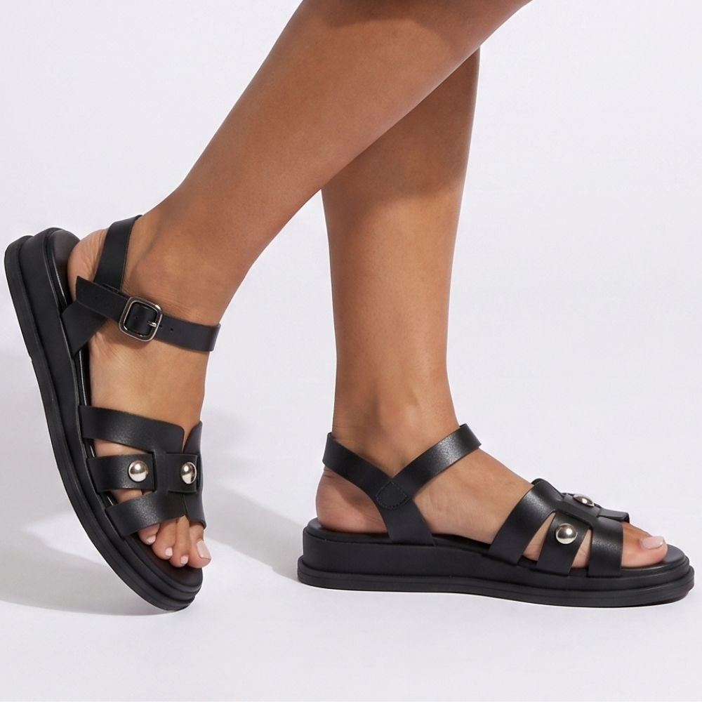 Black leather strappy sandals with buckle straps and decorative metal studs, shown on a model's feet against a light gray studio background.