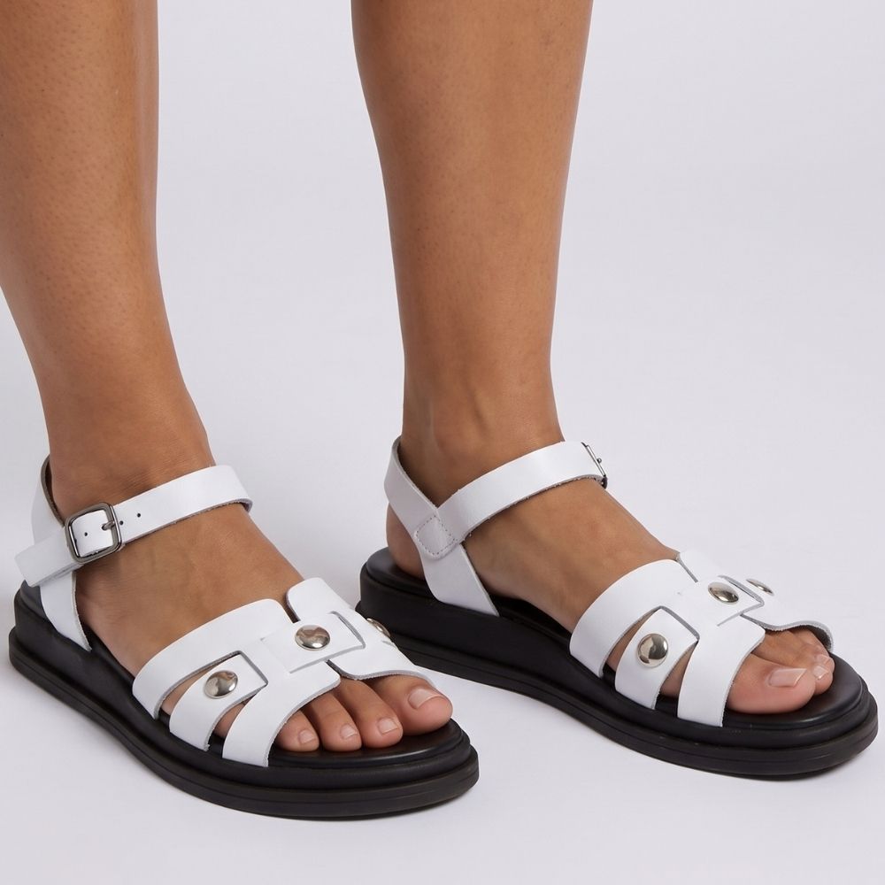 White leather strappy sandals with silver-tone hardware and black soles, shown on a model's feet against a light gray studio background.
